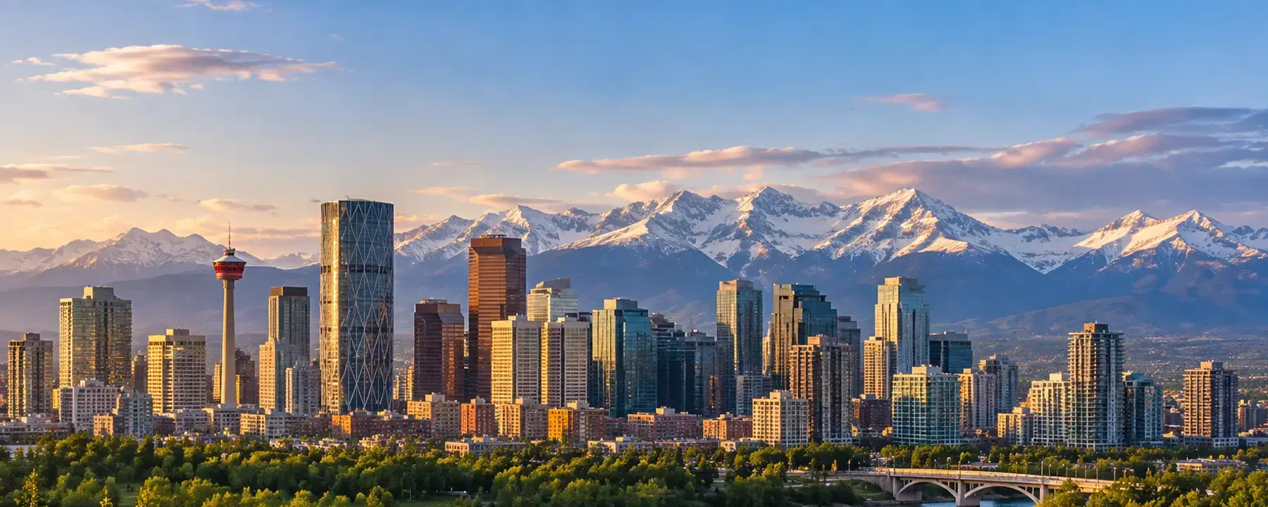 A city skyline at sunset with snow-capped mountains behind it and a river in front.